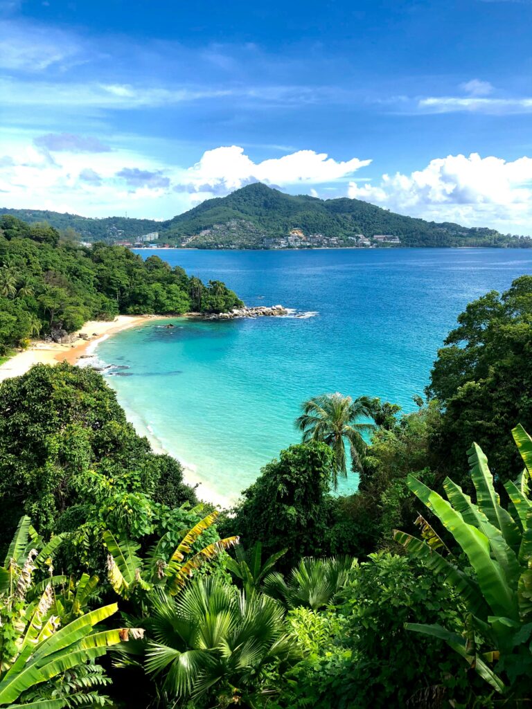 Udsigt over en tropisk ø nær Phuket i Thailand med turkisblåt vand, frodig skov, hvide sandstrande og skyer på himlen.