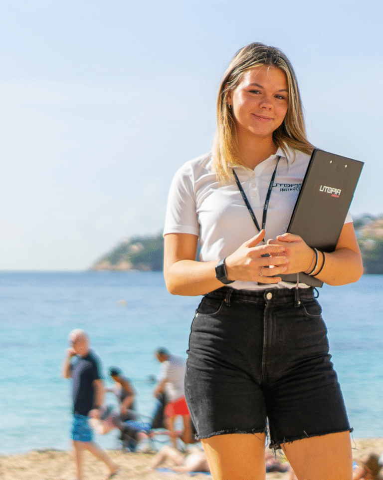 Ung kvinde i Utopia Guideskole-uniform med clipboard på stranden på Mallorca, smilende med hav og bjerge i baggrunden.